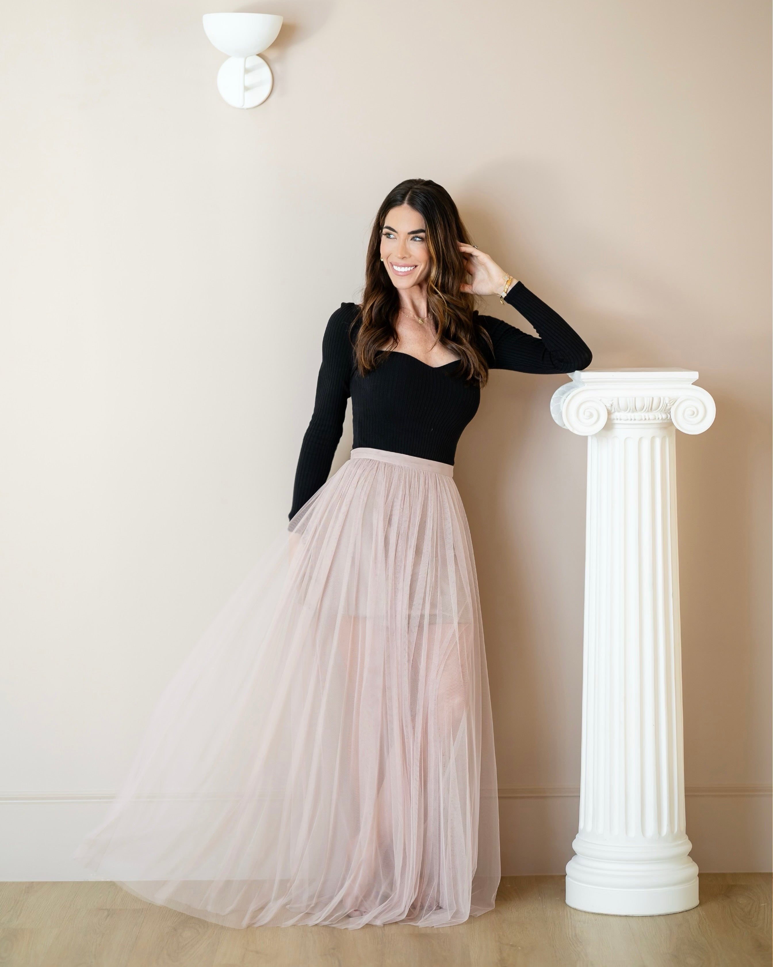 Caitlin Wilson in a black top and beige tulle skirt standing next to a white column against a beige wall.