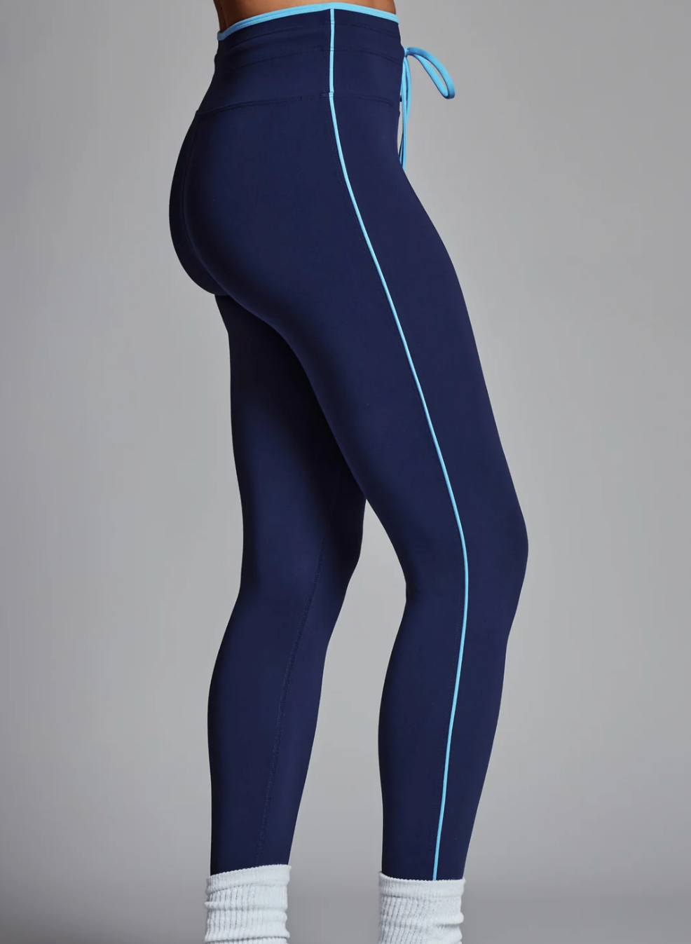 A side view of a person wearing navy blue high-waisted athletic leggings with a light blue side piping detail and matching drawstring, paired with white socks and sneakers against a solid gray studio background.